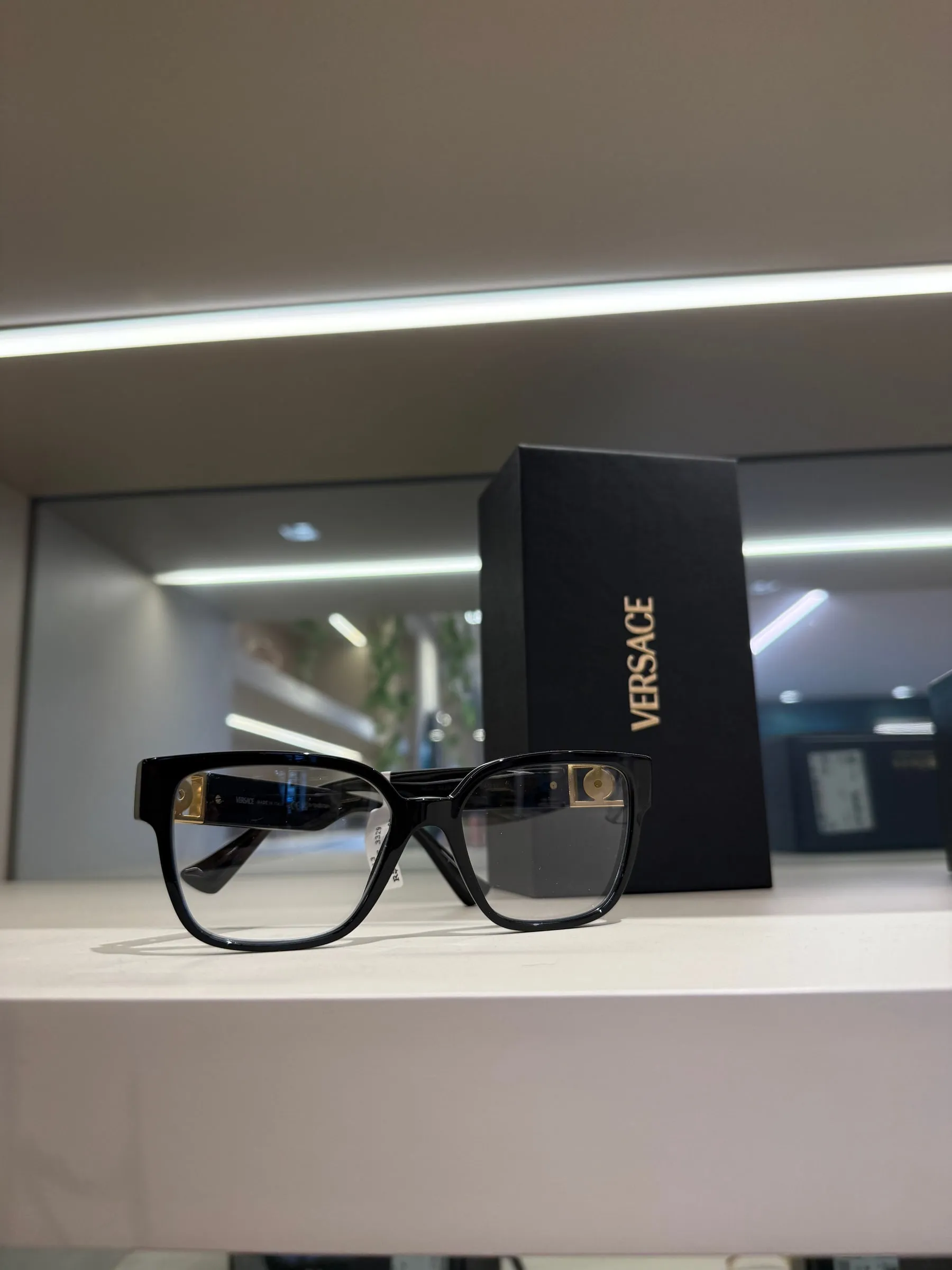 Armação de grau Versace preta com detalhes dourados, ao lado da caixa da marca